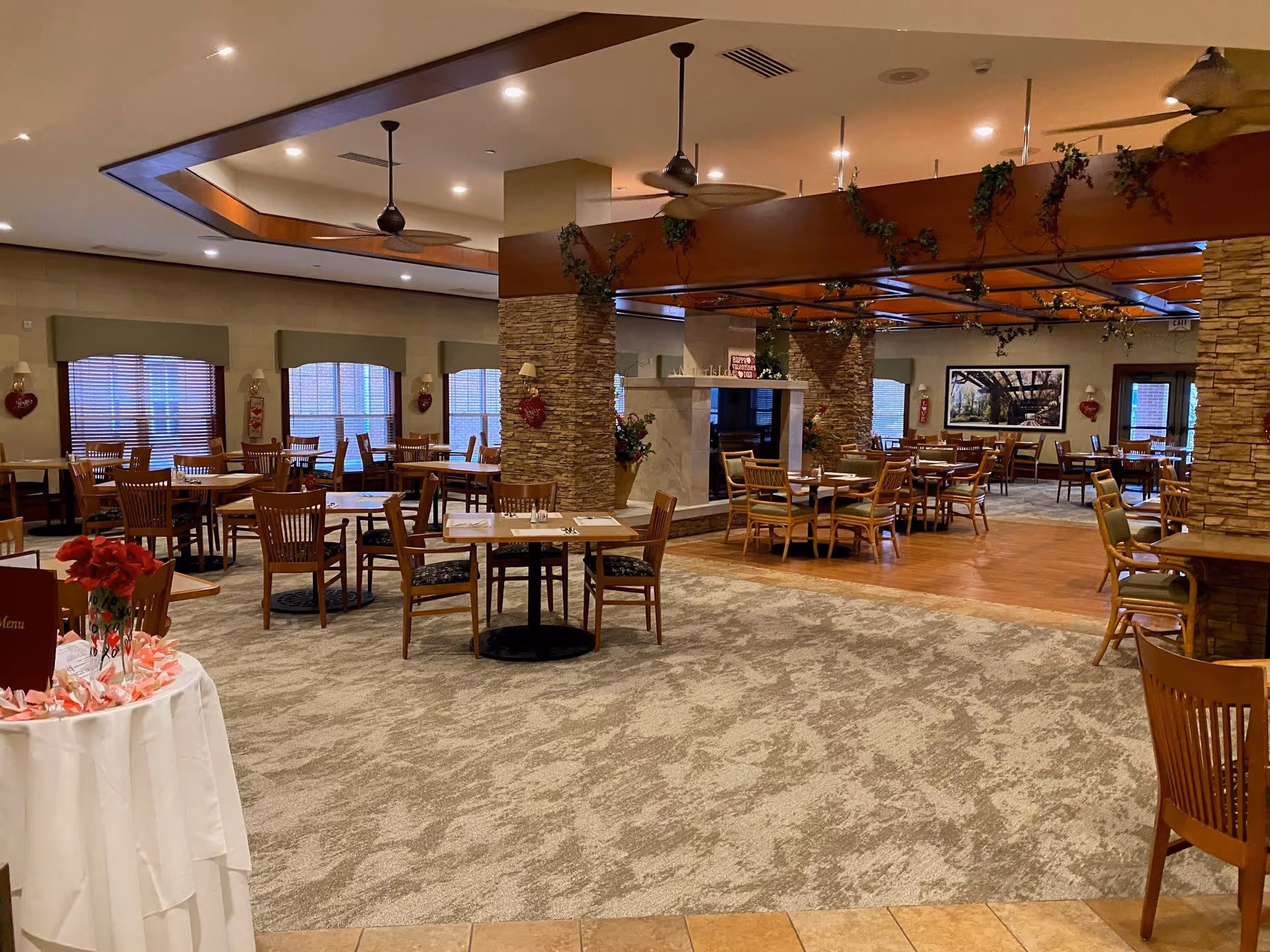 A spacious dining room with multiple wooden tables and chairs arranged neatly. The room features a carpeted floor with a section of wooden flooring near a stone pillar and a fireplace. Ceiling fans hang from the ceiling, and there are large windows with blinds along the walls. The room is decorated with plants and framed artwork, creating a warm and inviting atmosphere.