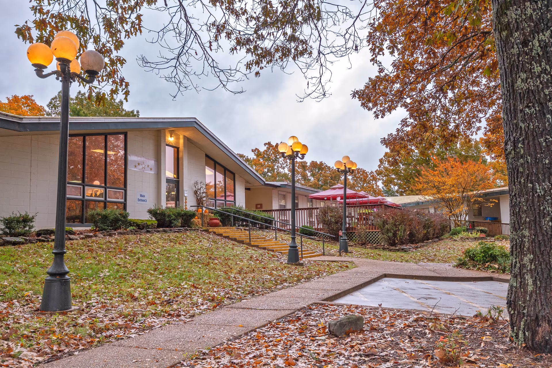Front exterior of a single-story nursing center with lamp posts, a sloped walkway and autumn trees.