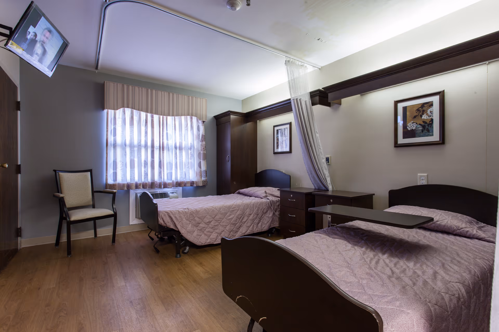 A shared bedroom in a healthcare facility with two single beds covered in light purple quilts. Each bed has a dark wooden headboard and a bedside table. A privacy curtain is partially drawn between the beds. There is a window with sheer curtains letting in natural light, a wooden chair near the window, and a wall-mounted TV in the corner. The room has wooden flooring and framed artwork on the walls.