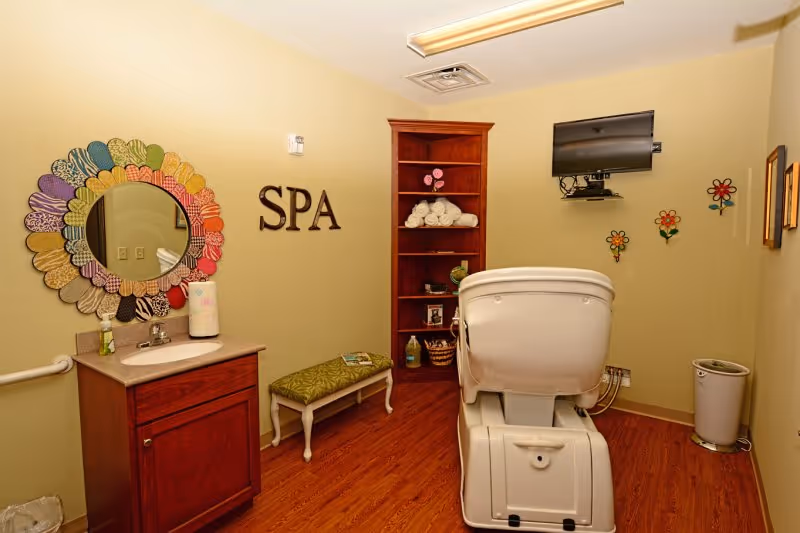 A spa room with a white spa chair facing a wall-mounted TV. The room has wooden flooring and light yellow walls. On the left side, there is a wooden cabinet with a sink and a colorful flower-shaped mirror above it. The word 'SPA' is mounted on the wall next to the mirror. A small bench with a green cushion is placed against the wall, and a tall wooden shelf holds rolled white towels and decorative items. Colorful flower wall decorations are on the wall near the TV.