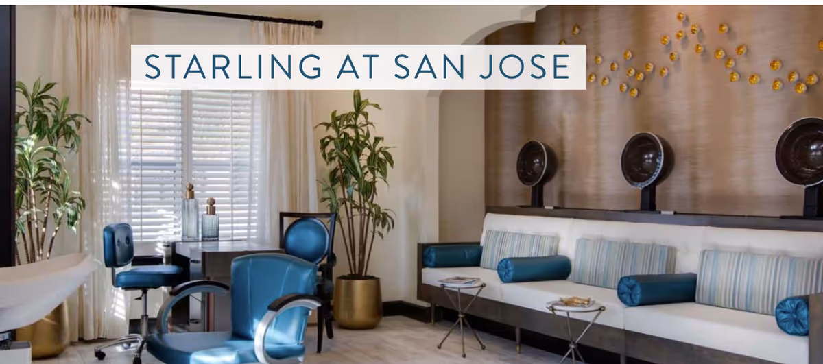 Interior view of a stylish salon area at Starling at San Jose featuring blue salon chairs, white cushioned bench seating with blue and striped pillows, hair drying stations, potted plants, and a window with white blinds and curtains.