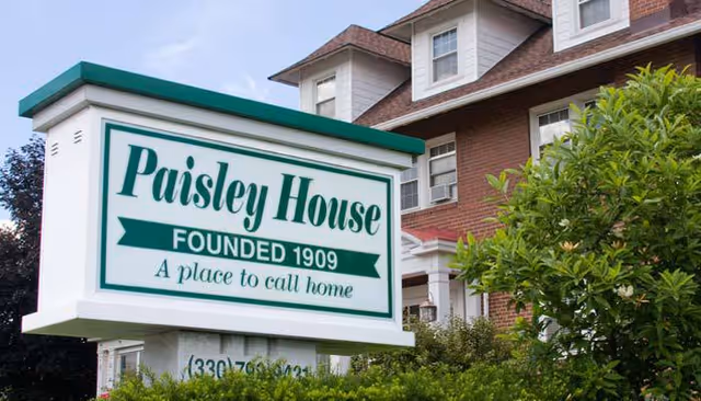A large white and green sign in front of a brick building reads 'Paisley House FOUNDED 1909 A place to call home'. The building has multiple windows and a sloped roof, surrounded by greenery and bushes.