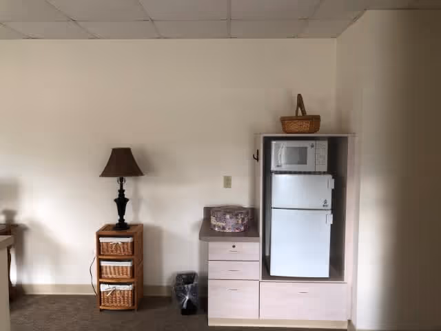 Interior view of a room with a small wooden cabinet holding a lamp and baskets on the left, and a light-colored cabinet on the right containing a microwave on top of a refrigerator, with a basket on top of the cabinet. There is a small trash bin on the floor between the two pieces of furniture.
