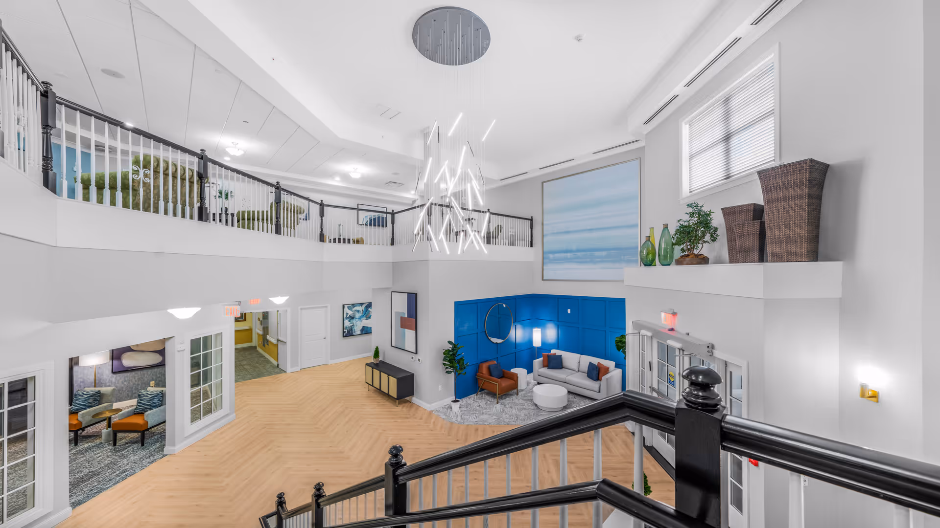 Two-story bright atrium lobby with a modern chandelier, seating area, and an upper balcony overlooking a staircase.