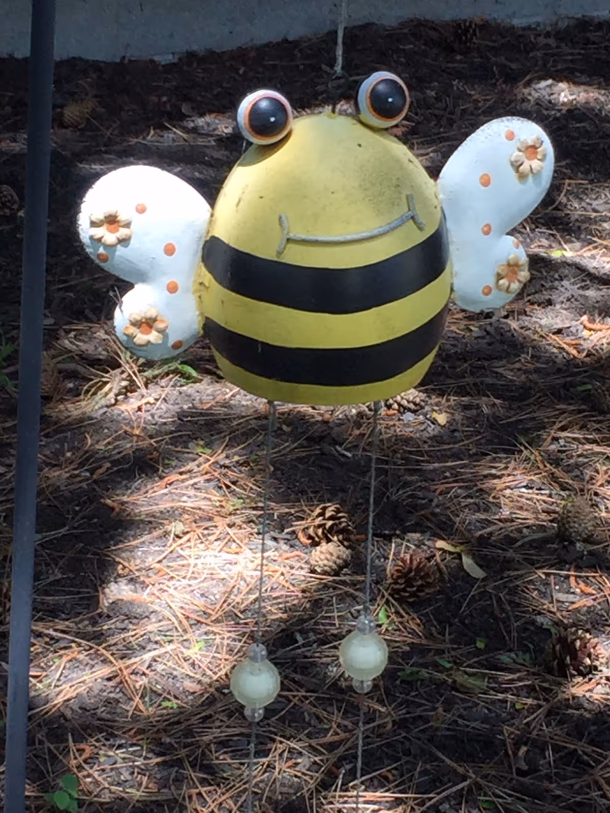 Whimsical metal bee garden ornament with yellow-and-black striped body, white flowered wings, and dangling beads set on pine-needle ground.