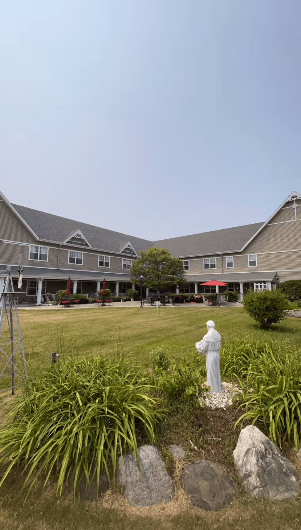 Outdoor courtyard area of a senior living facility with a large green lawn, a statue of a person in white robes surrounded by plants and rocks in the foreground, and a two-story building with beige siding and multiple windows in the background. There are red umbrellas and outdoor seating visible near the building.