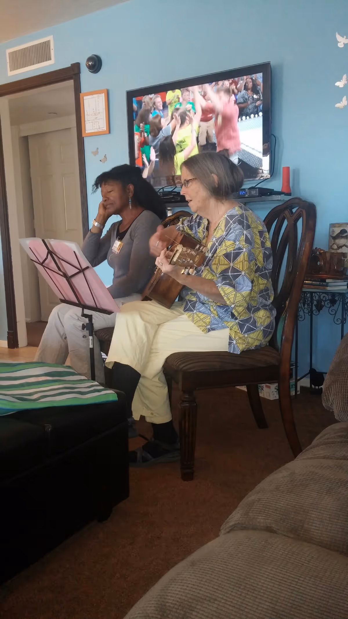 Two women sitting in a living room; one is playing a guitar while the other listens. A television mounted on a blue wall shows a group of people celebrating. The room has wooden chairs, a music stand with sheet music, and butterfly wall decorations.