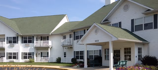 Exterior view of Sycamore Manor, a two-story white building with green roofs, multiple windows, balconies, and a covered entrance with the number 222 above it. There are benches and flower beds near the entrance.