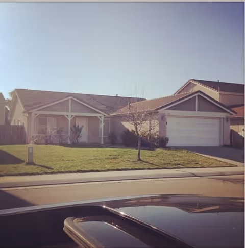 Single-story residential house with a front lawn, a small tree, and a two-car garage under a clear sky.