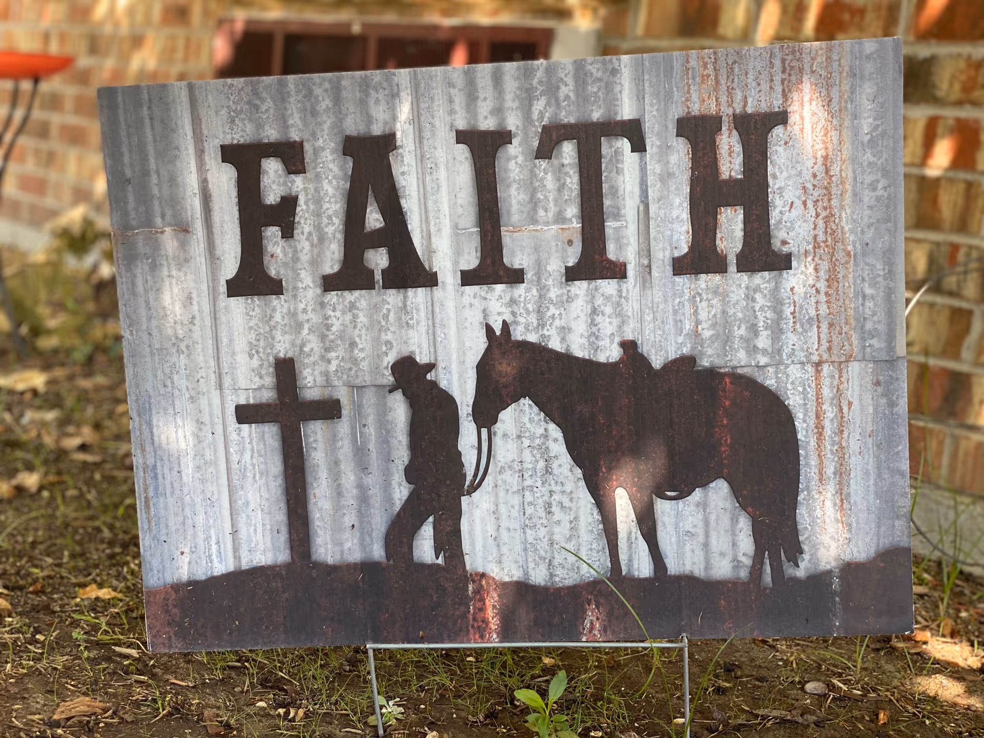 A metal sign reading "FAITH" with silhouettes of a person, a cross, and a horse displayed outdoors against a brick wall.