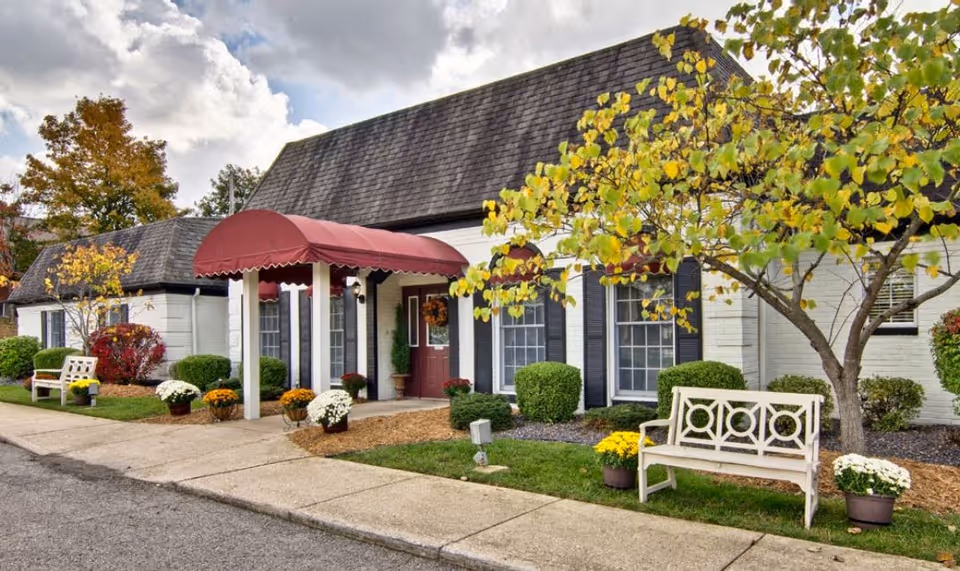 Exterior view of The Arbors at Parkside facility showing a single-story building with a dark shingled roof and white brick walls. The entrance has a red canopy and is decorated with potted flowers and shrubs. There are benches and small trees with autumn foliage along the sidewalk.