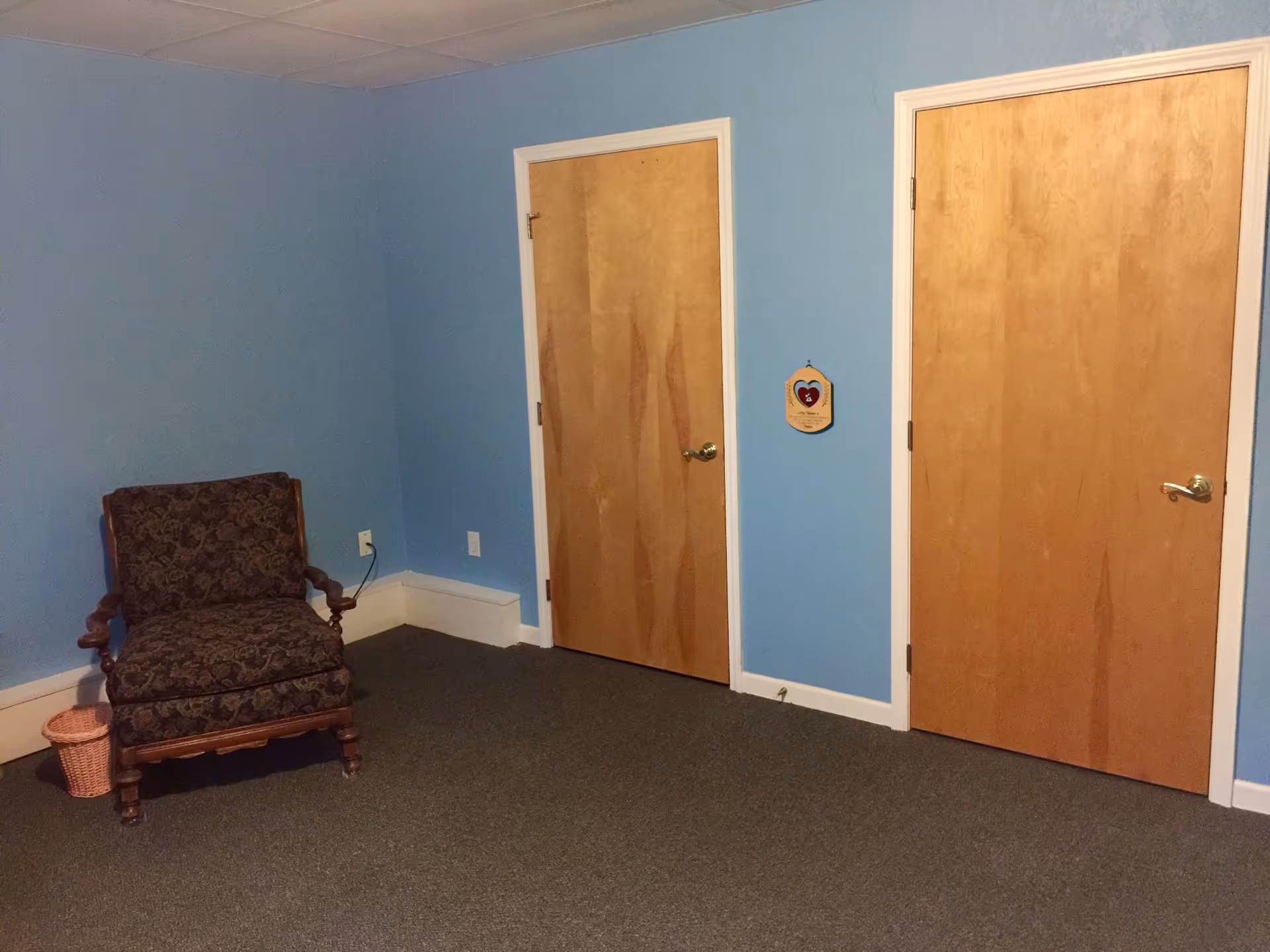 A room with light blue walls and gray carpeted floor featuring two closed wooden doors with silver handles. There is a dark patterned upholstered armchair with wooden arms and legs positioned near the corner, next to a small pink woven basket. A small decorative plaque with a heart design is hanging on the wall between the two doors.