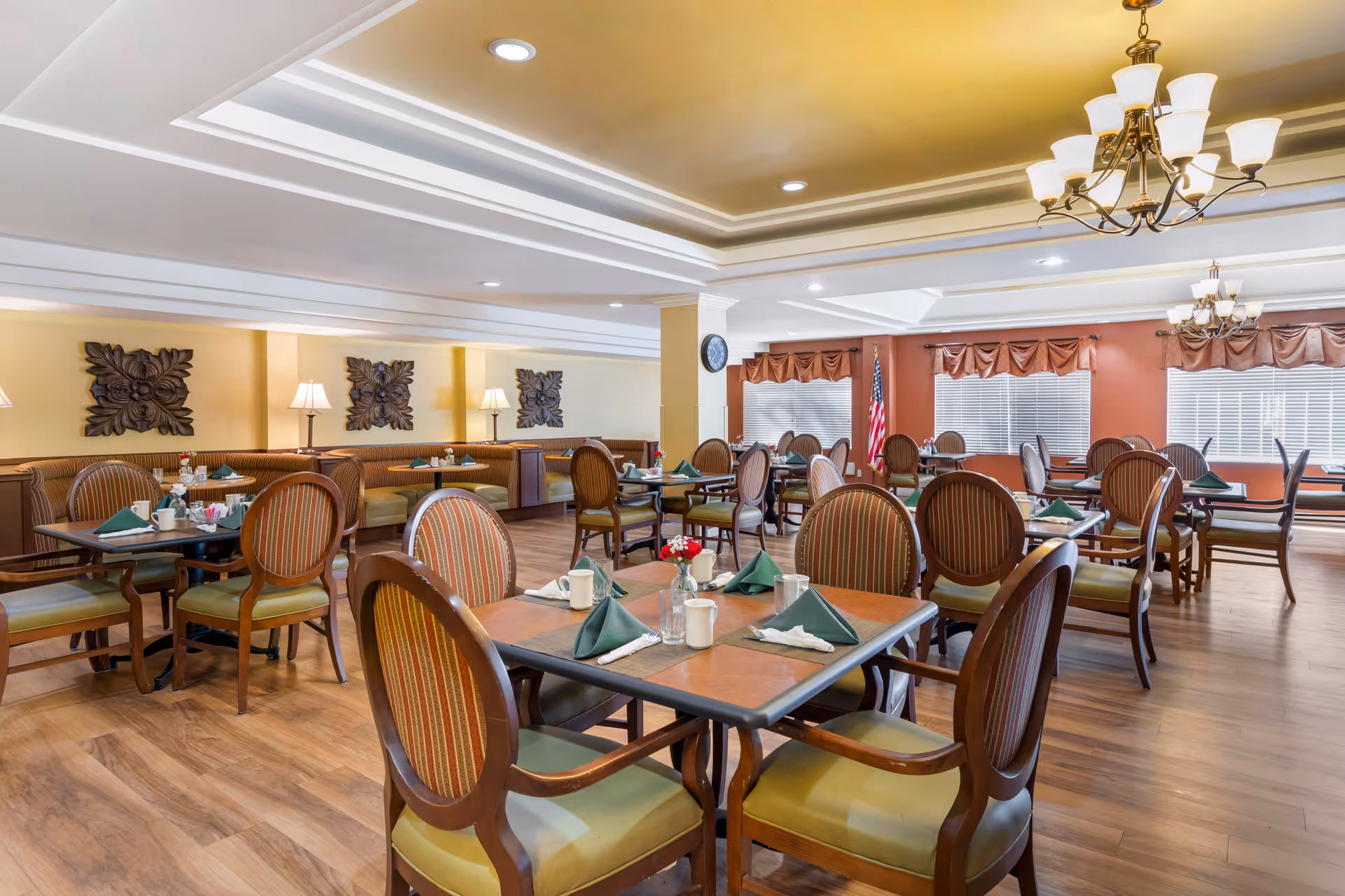 A spacious dining room with multiple wooden tables and chairs arranged neatly. Each table is set with green folded napkins, white mugs, and glassware. The room features warm lighting with ceiling chandeliers and recessed lights. There are decorative wall hangings and lamps along the walls, and large windows with blinds and valances. An American flag is visible near the windows.
