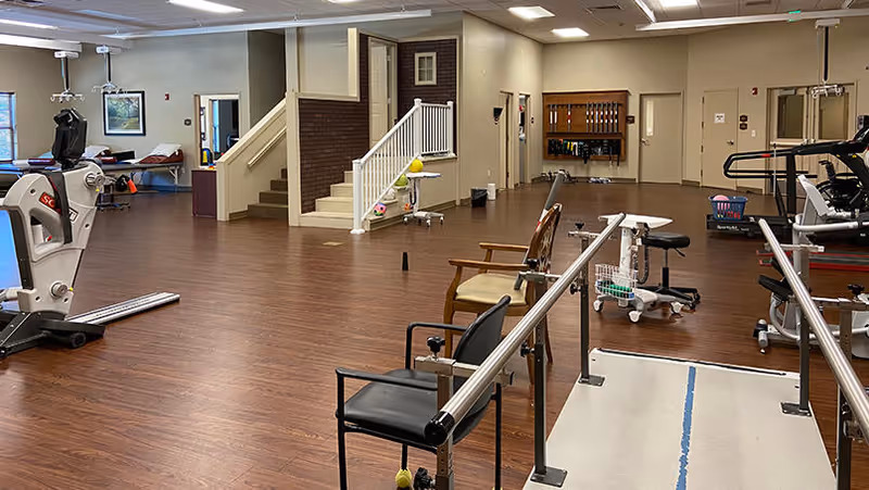 Interior view of a physical therapy and rehabilitation room with exercise equipment, parallel bars for walking practice, chairs, and a staircase. The room has wood flooring, beige walls, and fluorescent ceiling lights.