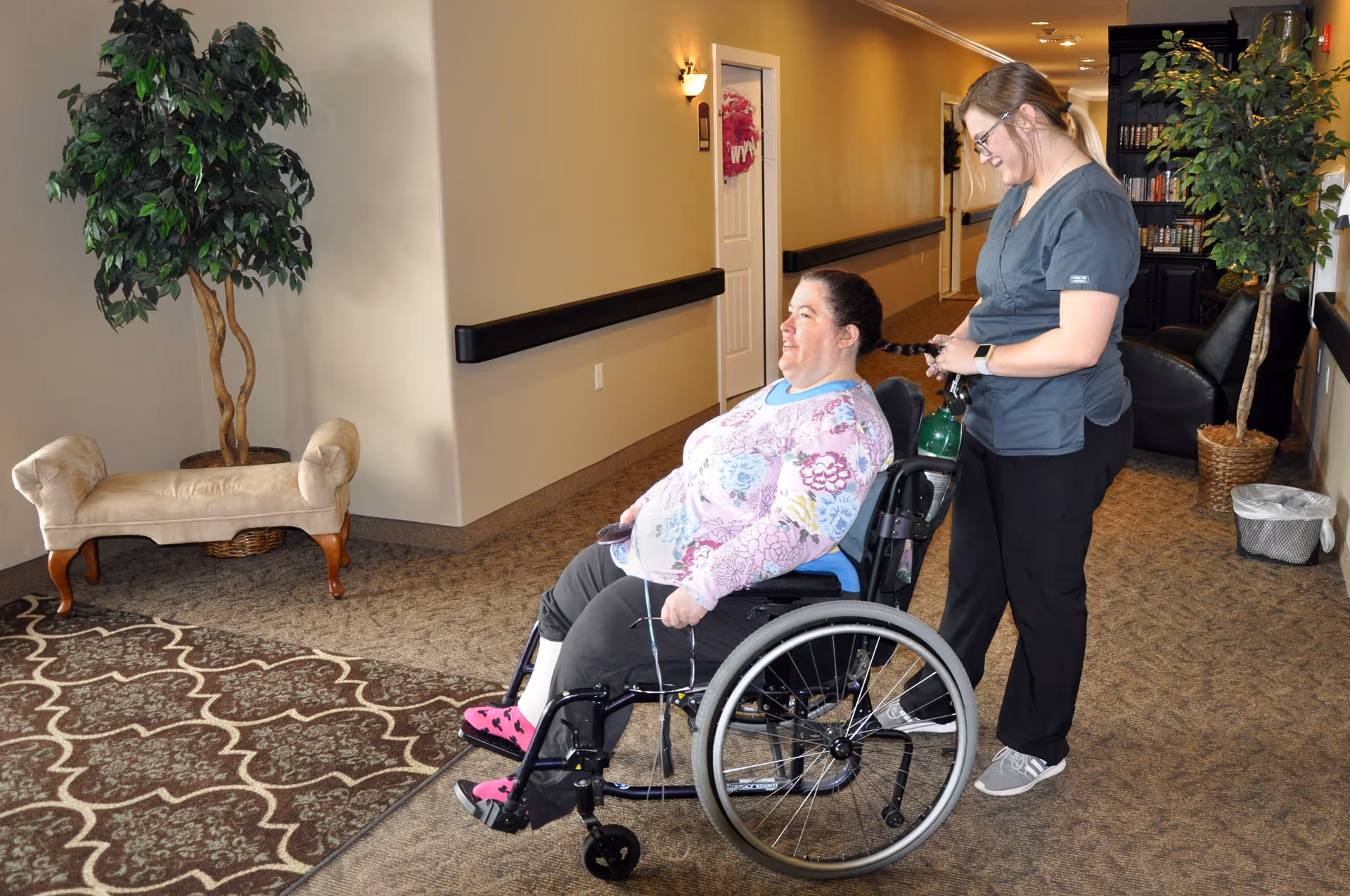 A caregiver standing behind a woman in a wheelchair in a hallway of a senior living facility. The caregiver is smiling and holding the woman's braided hair. The hallway has carpeted floors, a potted plant, a small upholstered bench, and a bookshelf with chairs in the background.