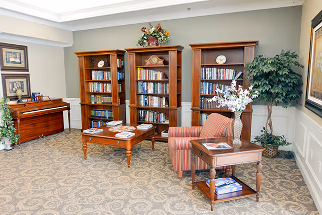 A cozy reading room with three wooden bookshelves filled with books, a wooden coffee table with magazines, a striped armchair, a side table with a vase of flowers, a piano in the corner, and decorative plants and framed artwork on the walls.