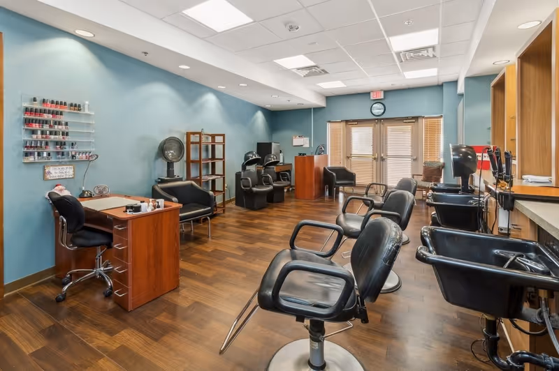 Interior view of a salon area in a senior living facility with multiple black salon chairs, hair washing stations, a manicure table with nail polish on the wall, and wooden flooring. The room has blue walls, overhead lighting, and a double door with blinds at the far end.