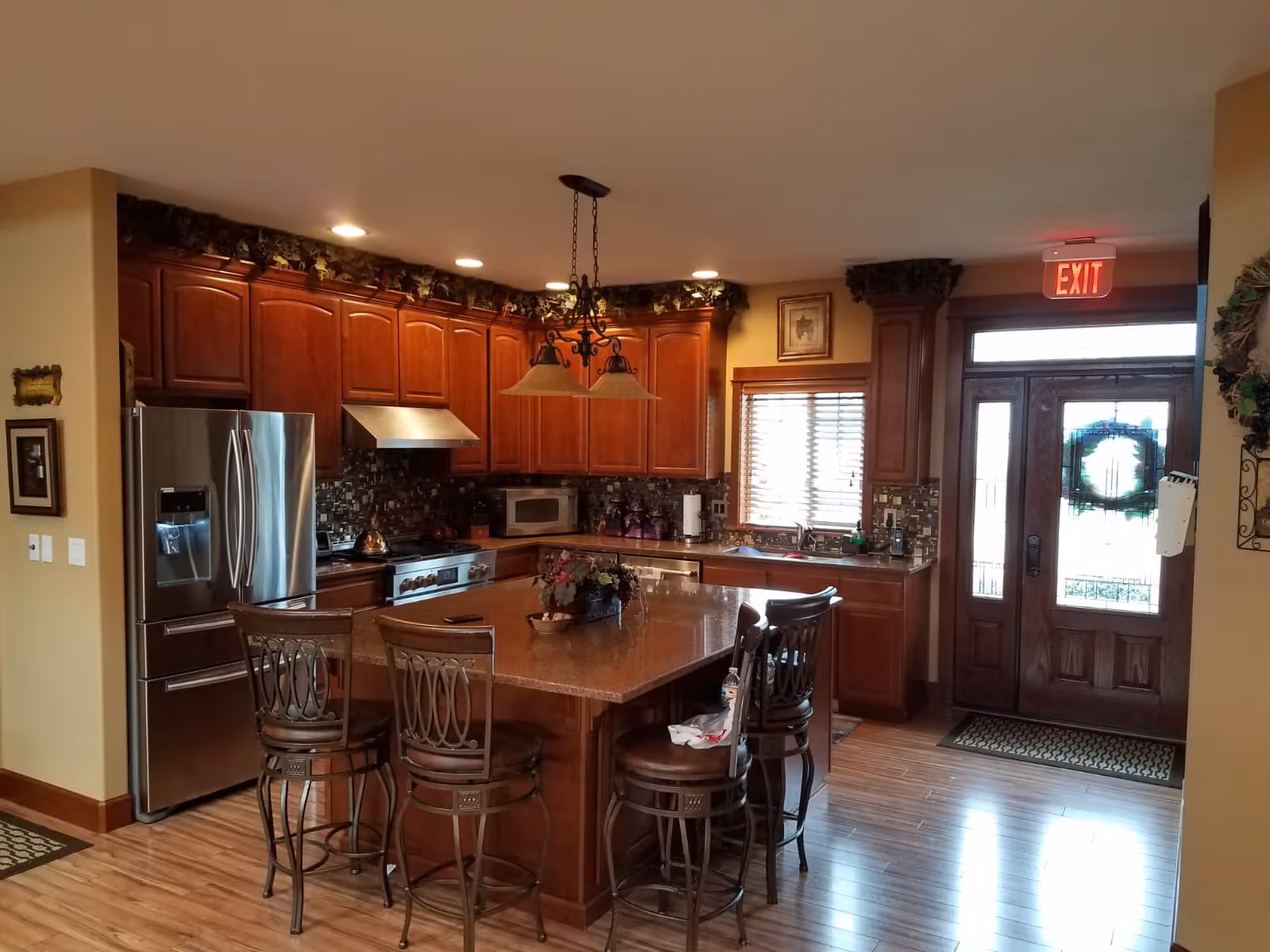 Interior view of a kitchen with wooden cabinets, a stainless steel refrigerator, stove, microwave, and a large island with four high chairs. The kitchen has a tiled backsplash and wooden flooring. There is a window with blinds above the sink and a wooden door with glass panels and a wreath. An exit sign is visible above the door.