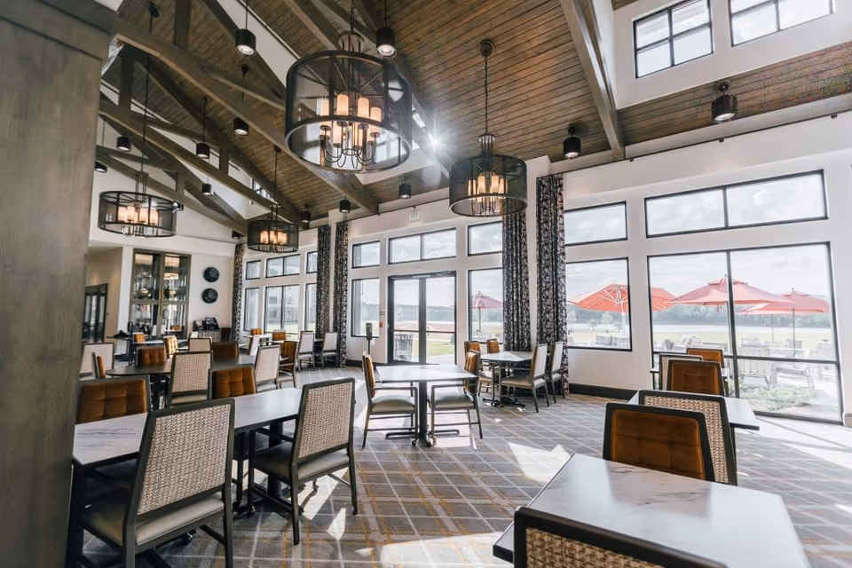 Bright dining room with multiple tables and chairs arranged neatly. Large windows and glass doors allow natural light to fill the space, showing a view of outdoor seating with red umbrellas. The ceiling features wooden beams and modern hanging light fixtures.