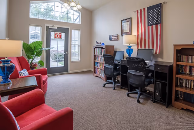 A bright room with a glass door marked 'NOT AN EXIT' and large windows letting in natural light. The room features two red armchairs with a side table and blue lamp, two desks with computers and office chairs, bookshelves filled with books, and an American flag hanging on the wall.