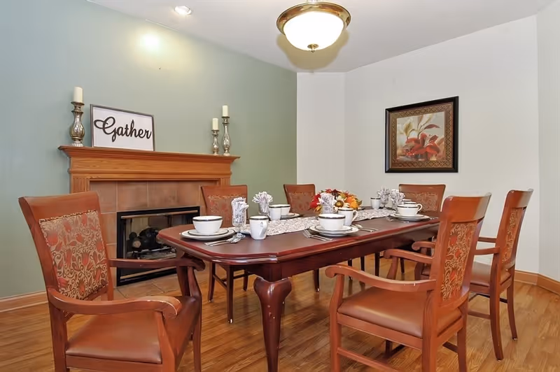 A dining room with a wooden table set for six people with white cups, plates, and napkins. The room features six wooden chairs with patterned upholstery, a fireplace with a wooden mantle, and a decorative sign that says 'Gather'. There is a framed floral painting on the wall and a ceiling light fixture.