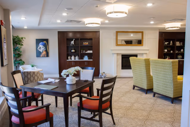 A cozy senior living common area with a dark wooden table surrounded by four chairs with red cushions. The room features a fireplace with a gold-framed mirror above it, two green armchairs facing the fireplace, built-in dark wood shelves with decorative items, a potted plant, and artwork on the walls. The carpet has a subtle pattern and the ceiling has recessed lighting and modern circular light fixtures.