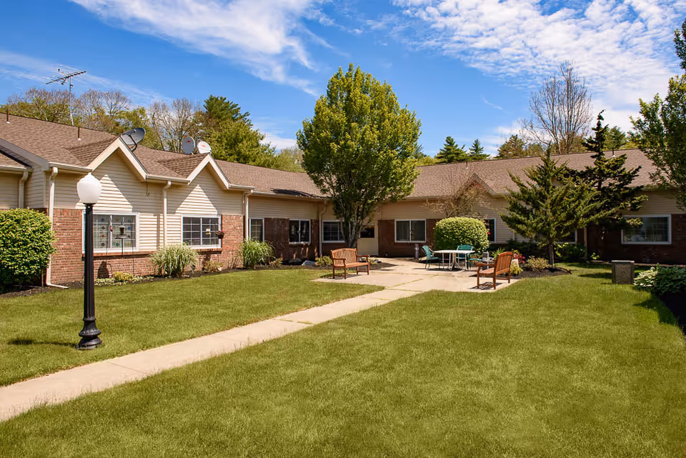 Sunny courtyard of a single-story senior living building with a lawn, walkway, benches, tables, and trees.