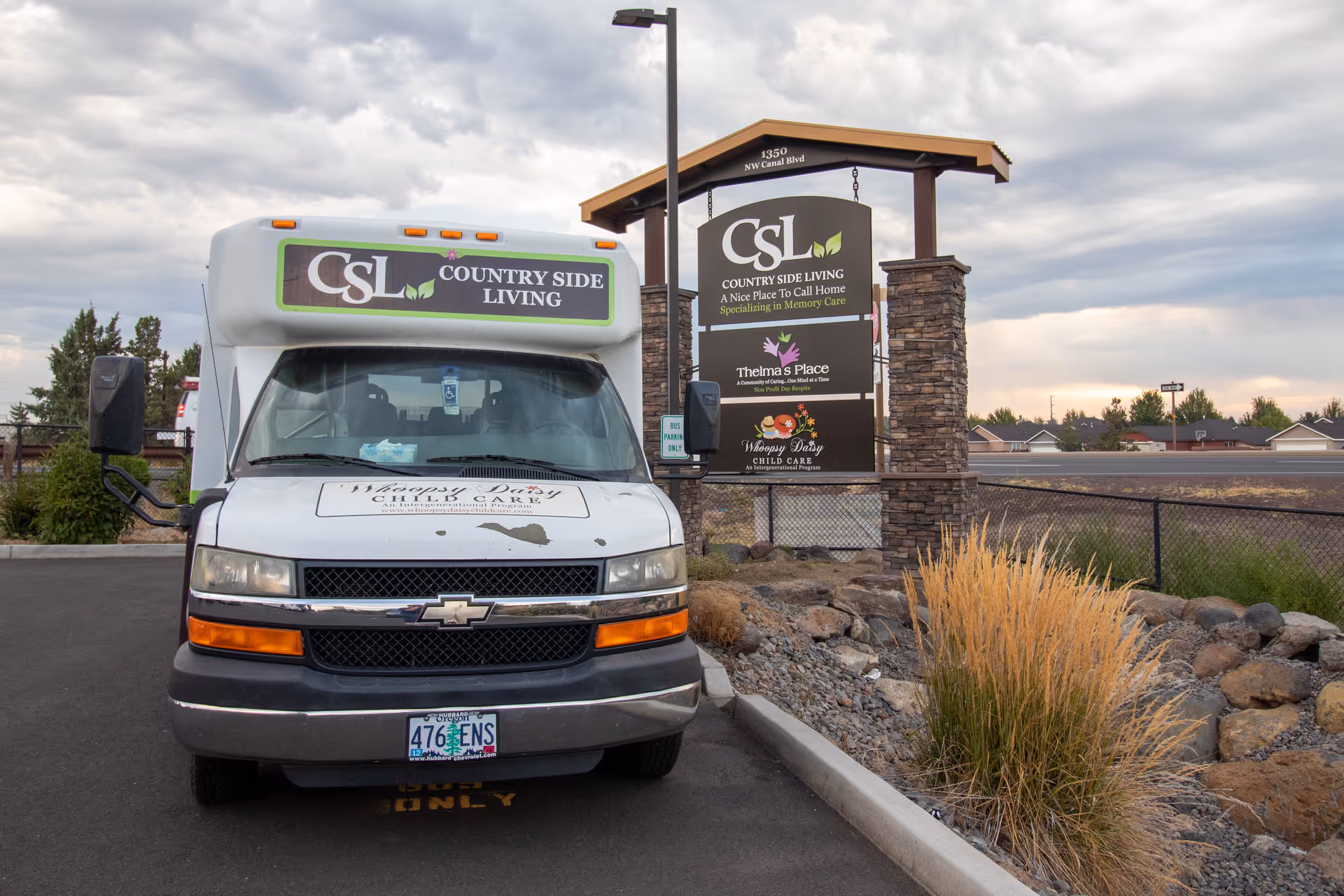 A white shuttle van with the sign 'CSL Country Side Living' on the front is parked in a lot near a stone and wood signpost. The signpost displays 'CSL Country Side Living, A Nice Place To Call Home, Specializing in Memory Care', along with additional signs for 'Thelma's Place' and 'Whoopsy Daisy Child Care'. The background shows a cloudy sky and some residential houses.