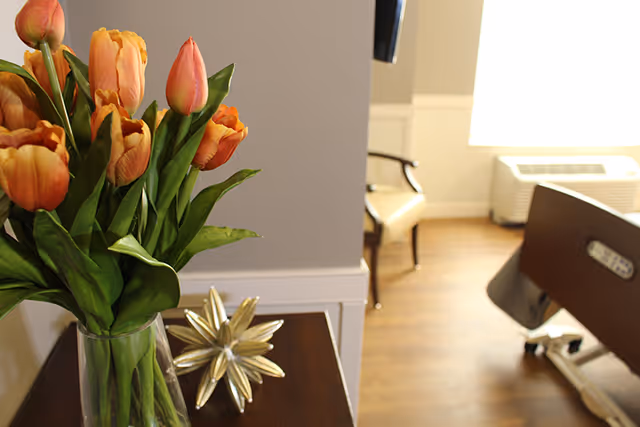A vase of orange tulips on a bedside table with a decorative star and a partial view of a hospital-style bed and chair in a senior care room.