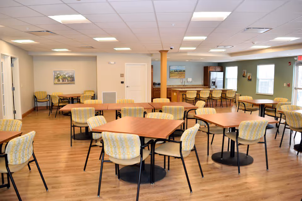 A spacious dining room with multiple wooden tables and cushioned chairs arranged neatly. The room has a light wood floor, beige and green walls, and a ceiling with recessed lighting. There is a kitchen area with a counter and bar stools in the background, along with windows letting in natural light and some wall decorations.