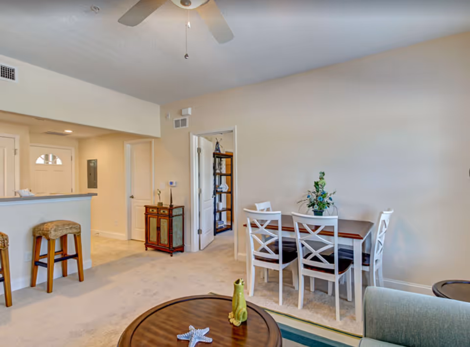 Open-plan living area with a round coffee table in the foreground, a sofa, bar stools at a counter, and a dining table with four chairs against a beige wall.