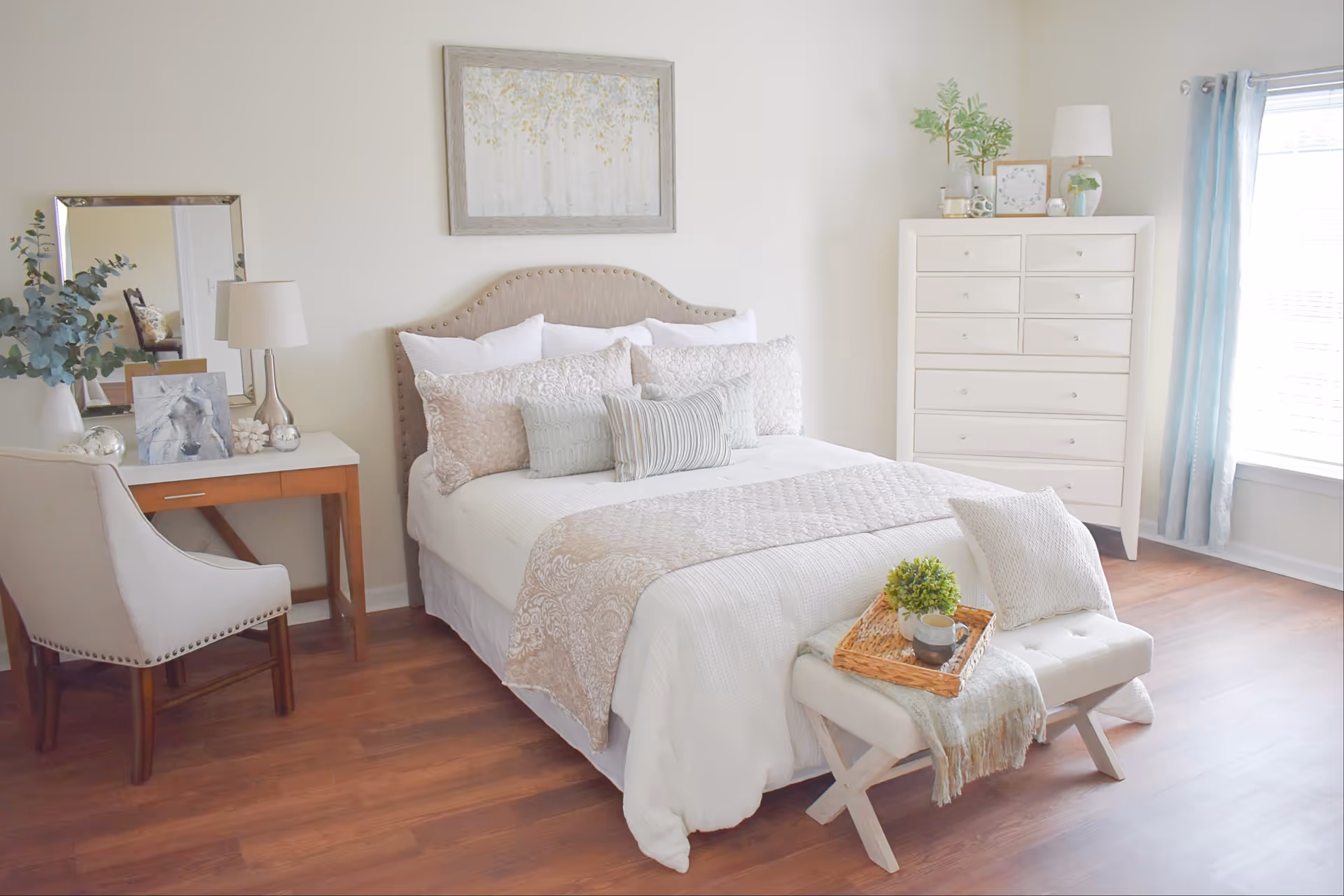 A bright and cozy bedroom featuring a neatly made bed with multiple pillows and a beige patterned throw. At the foot of the bed is a cushioned bench with a tray holding a small plant and a cup. To the left, there is a wooden desk with a chair, a table lamp, a mirror, and decorative items. On the right side of the room, a white dresser stands near a window with light blue curtains, allowing natural light to fill the space. The room has wooden flooring and light-colored walls, creating a warm and inviting atmosphere.