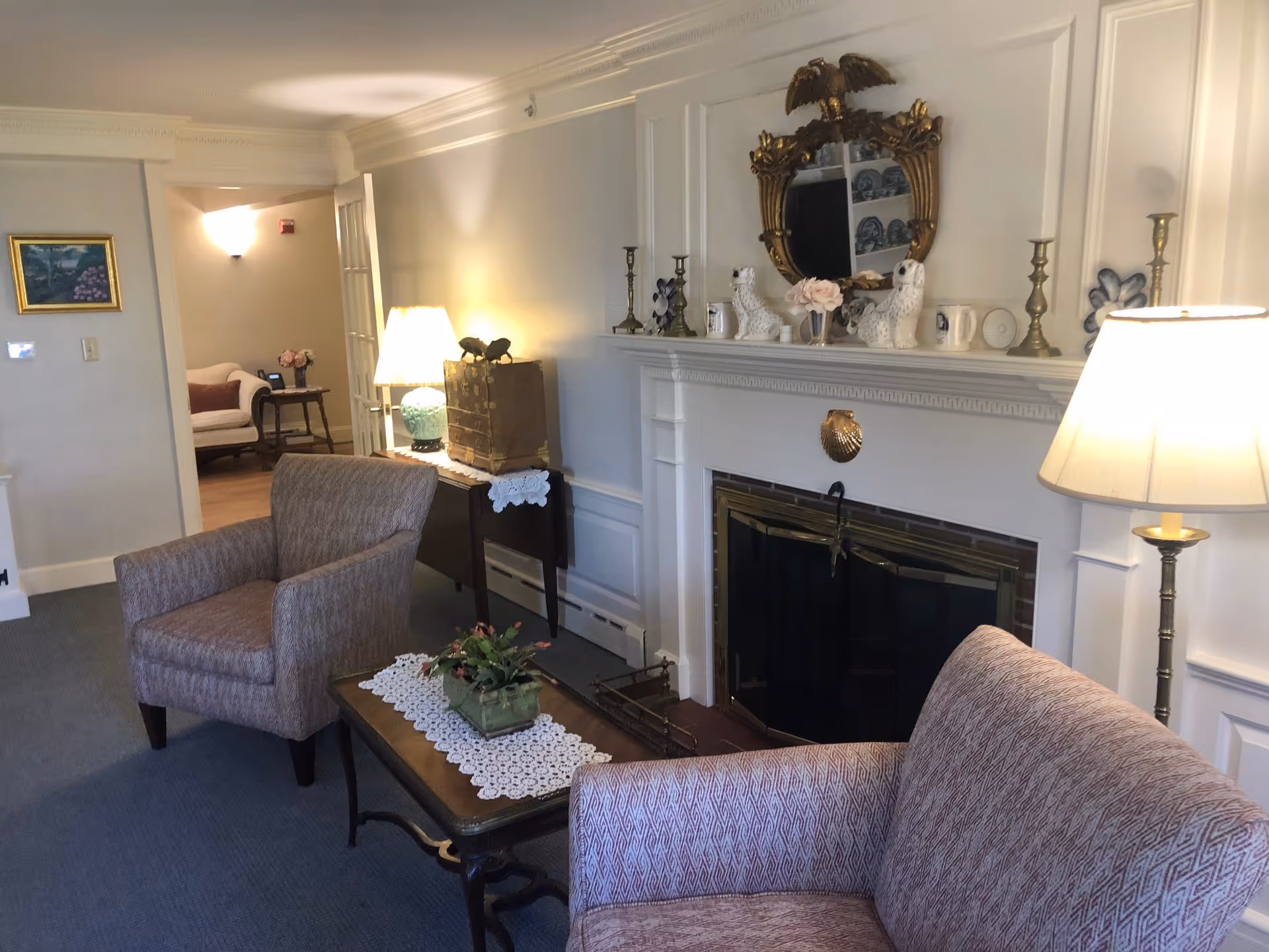 A cozy living room area with two upholstered armchairs and a small wooden coffee table with a lace runner and a potted plant. There is a white fireplace with a decorative gold-framed mirror above it, flanked by candlesticks and ceramic dog figurines. A floor lamp and a table lamp provide warm lighting. In the background, there is a glimpse of another room with a sofa and a side table with flowers.