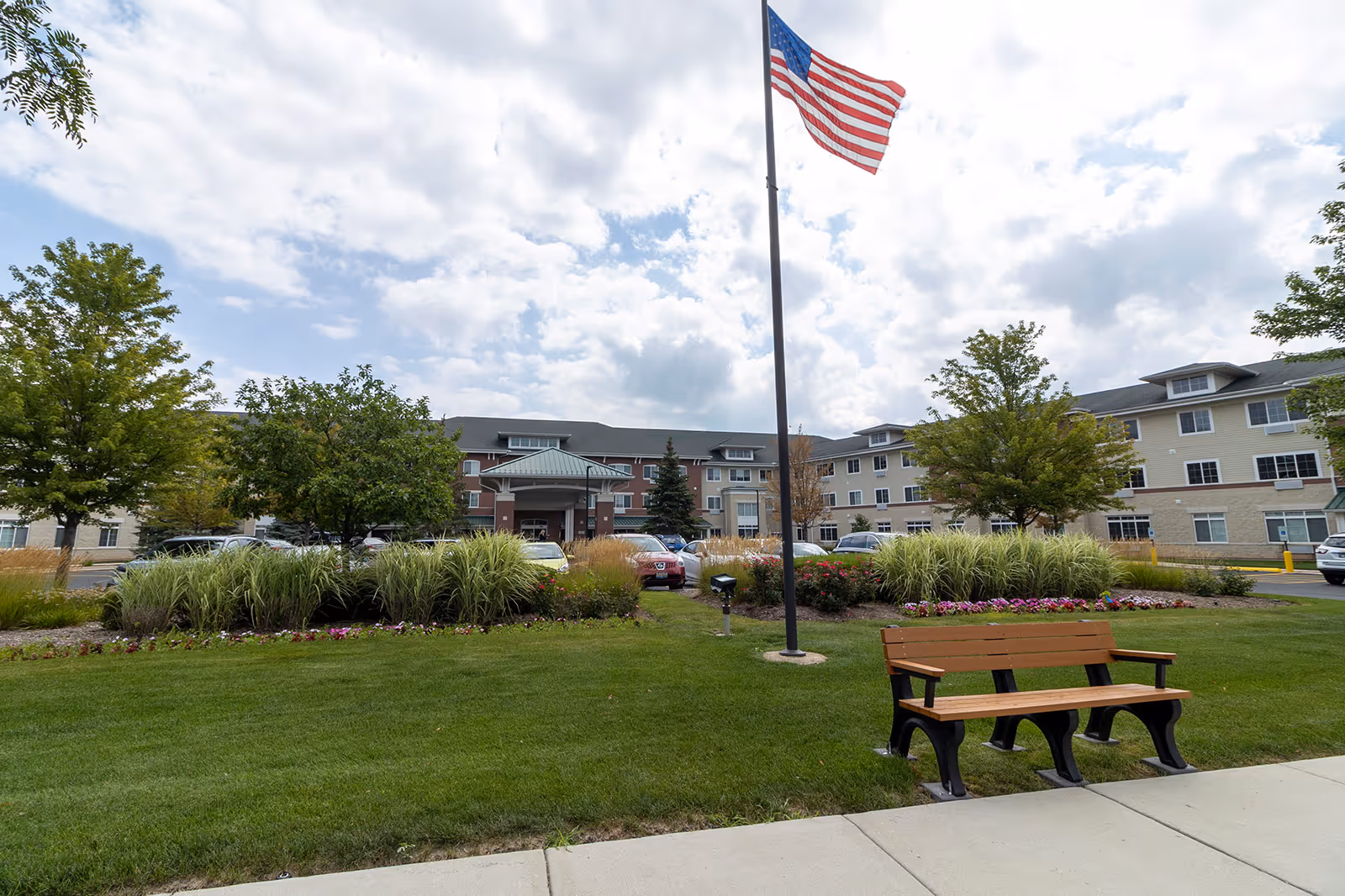 Front exterior of a multi-story senior living building with landscaped lawn, an American flag on a flagpole, and a bench in the foreground.