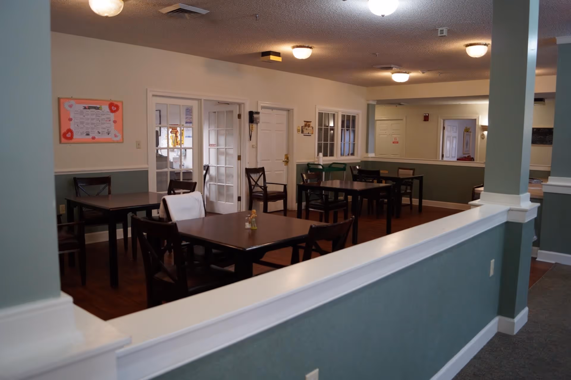 Interior view of a dining area in a senior living facility with several dark wooden tables and chairs arranged neatly. The walls are painted in a two-tone color scheme with light green on the lower half and white on the upper half. There are multiple ceiling lights providing illumination, and a bulletin board with decorations is visible on one wall. Glass-paneled doors and white doors lead to other rooms.