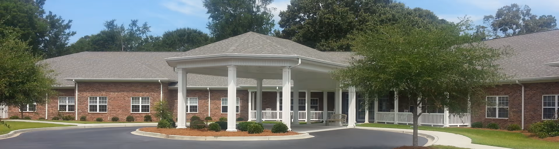 Front exterior view of a single-story brick building with a covered entrance supported by white columns, surrounded by trees and a paved driveway.