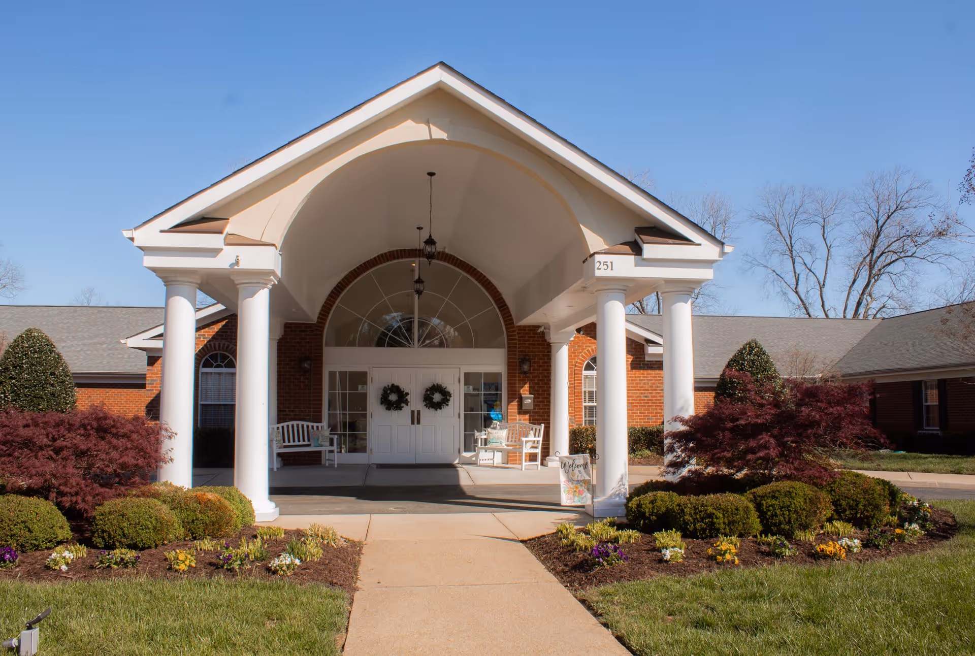 Front exterior view of a senior living facility with a covered entrance supported by white columns. The building is made of red brick with white doors decorated with wreaths. There are benches on either side of the entrance and well-maintained landscaping with bushes and flowers in front.