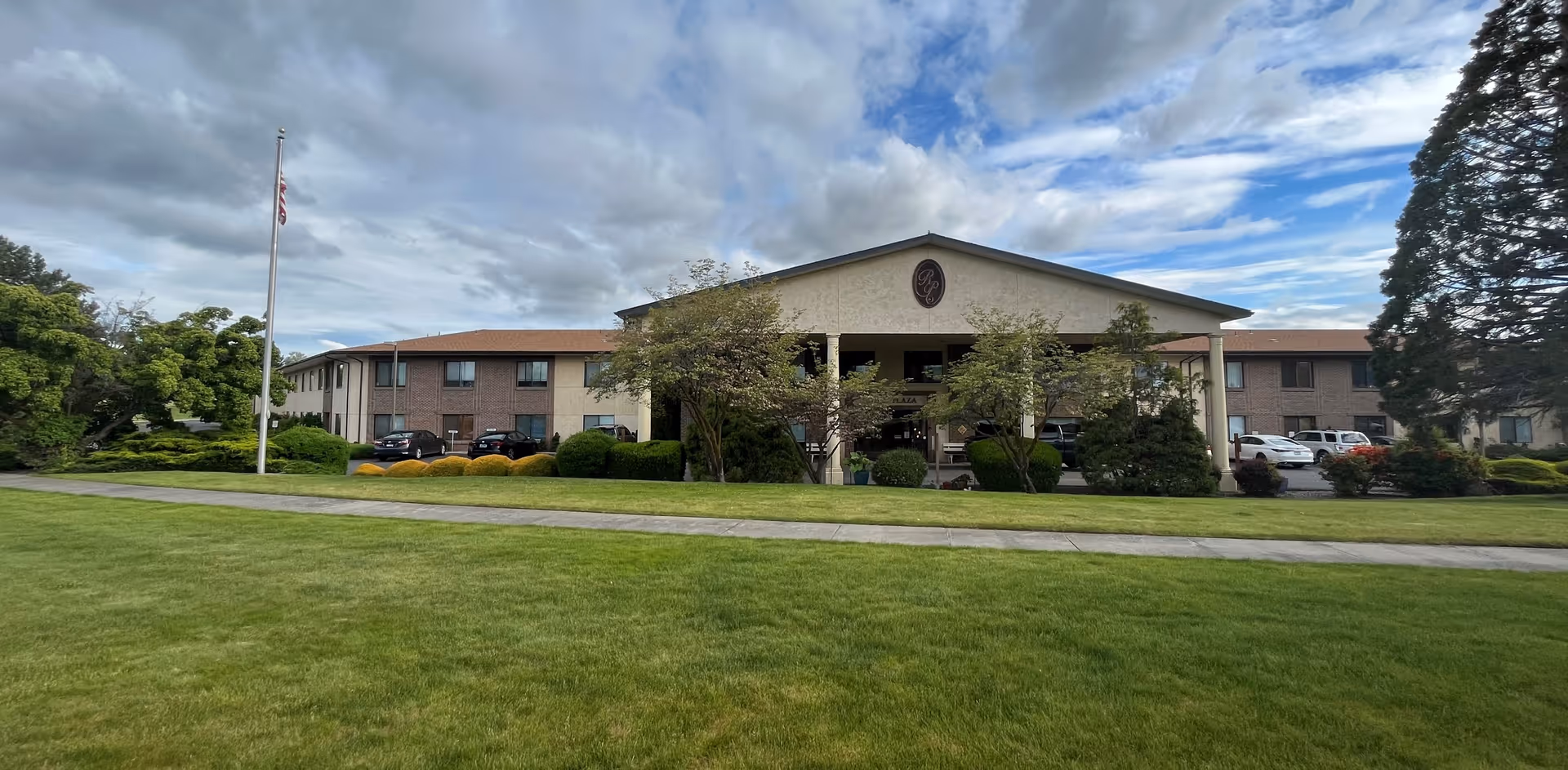 Front exterior view of a two-story retirement living facility named Royal Plaza of Olympus Retirement Living, with a well-maintained lawn, trees, bushes, a flagpole with an American flag, and a partly cloudy sky.