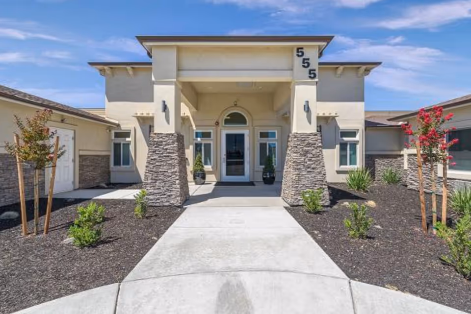 Front exterior view of a modern single-story building with beige walls, stone pillars, and a covered entrance. The building has the number 555 displayed vertically on the right pillar. There are small landscaped areas with young trees and shrubs on either side of the concrete walkway leading to the entrance.