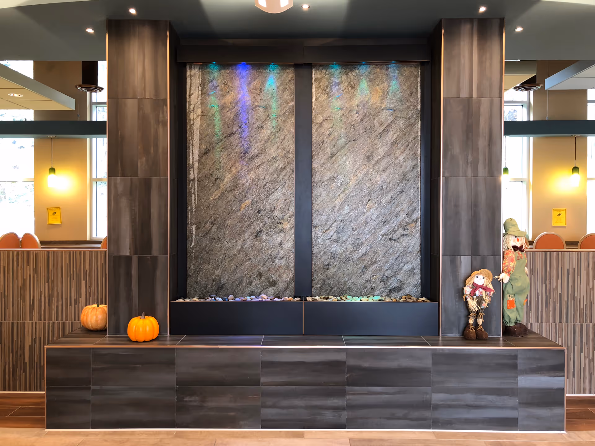 Indoor lobby water feature with a large stone wall, tiled bench, colorful lights, pumpkins and decorative scarecrow dolls on the ledge.