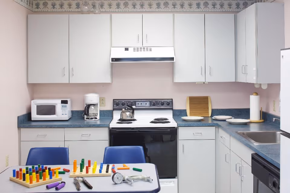 A small kitchen with white cabinets and blue countertops. The kitchen includes a microwave, coffee maker, stove with a kettle, sink, and dishwasher. On the table in the foreground, there are colorful pegboard games and some utensils. The walls are light pink with a floral wallpaper border near the ceiling.