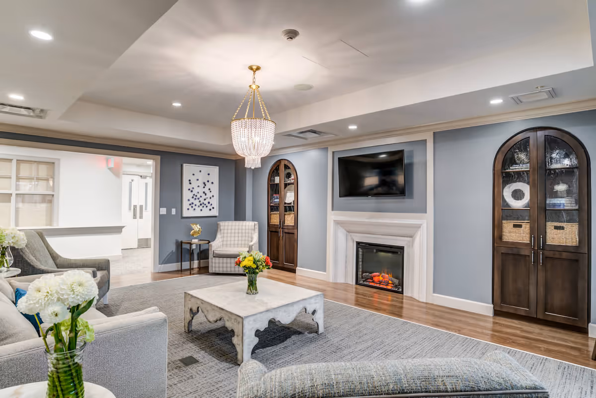 A well-appointed common living room with sofas, a central coffee table, a fireplace beneath a wall-mounted TV, built-in cabinets, and a chandelier.