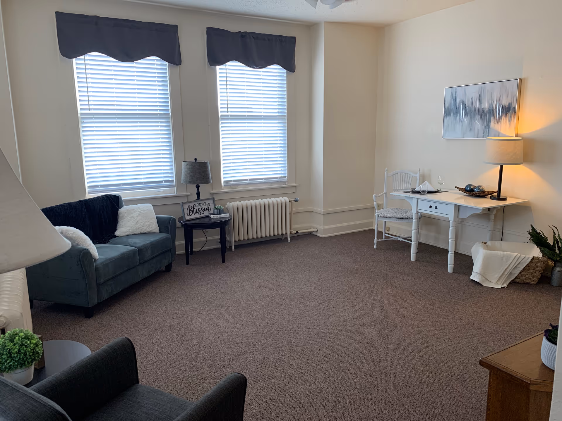 Cozy living room in a senior living facility with sofa, desk, chairs, and large windows.