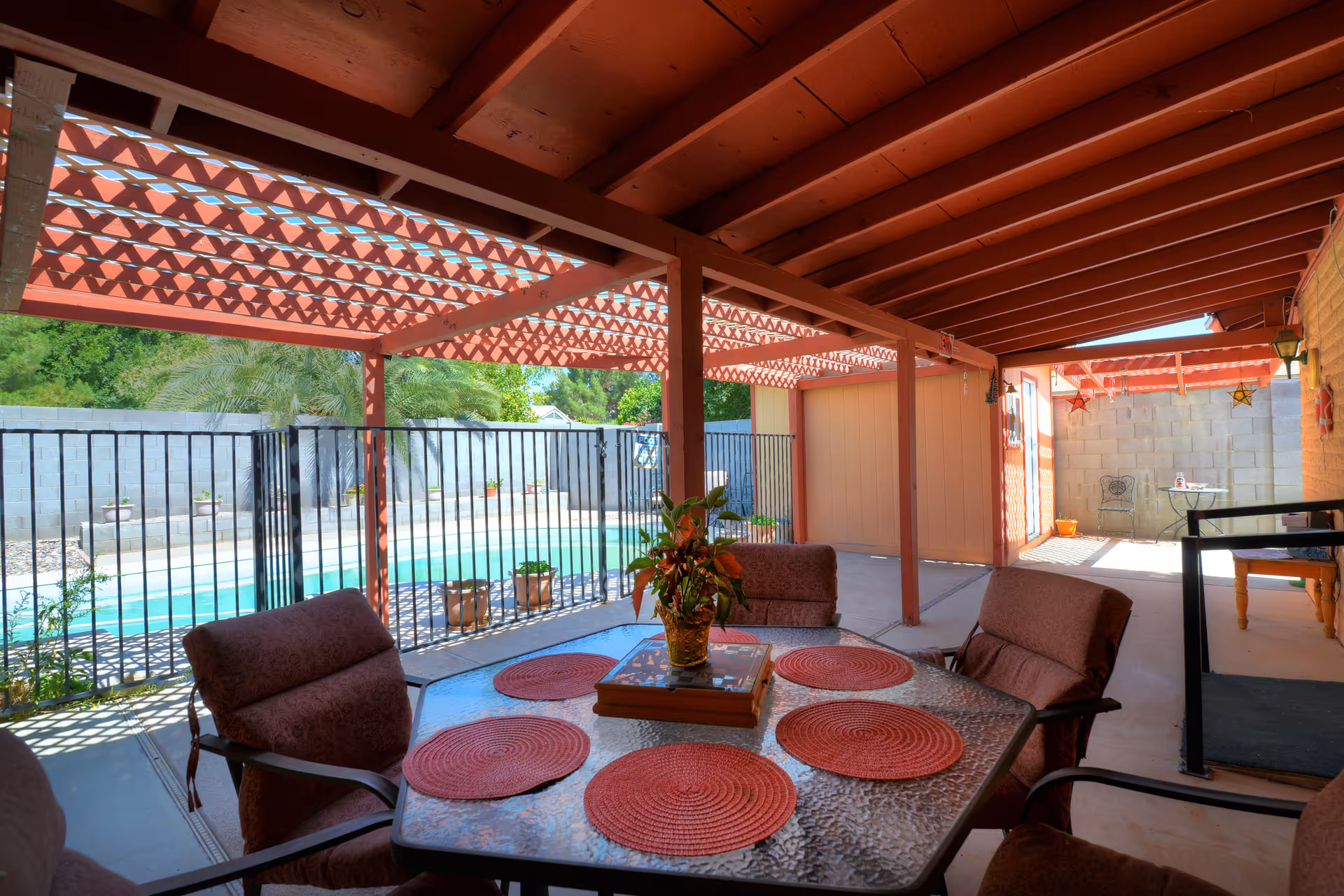 Covered outdoor patio area with a glass table set with red placemats and cushioned chairs. The patio has a red wooden pergola roof and overlooks a fenced swimming pool with greenery and a concrete block wall in the background.