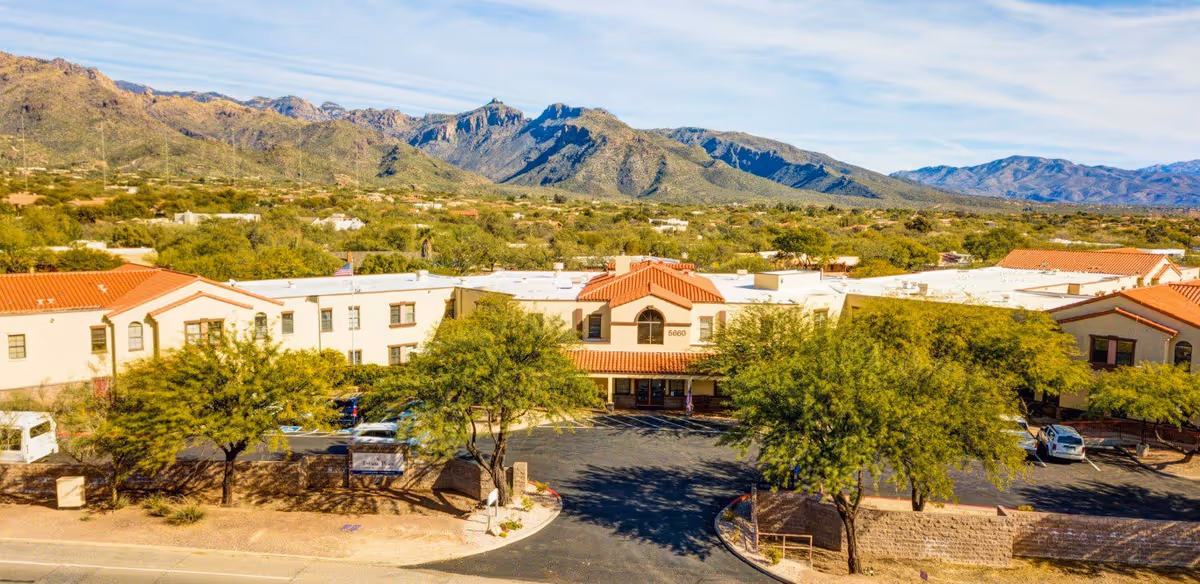 Aerial view of Tucson Place at Ventana Canyon, a senior living facility with a beige and terracotta roof building surrounded by trees and parking spaces, set against a backdrop of mountains under a partly cloudy sky.