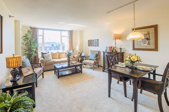 Bright, carpeted living room with a beige sofa, coffee table, dining table, and large window letting in natural light.