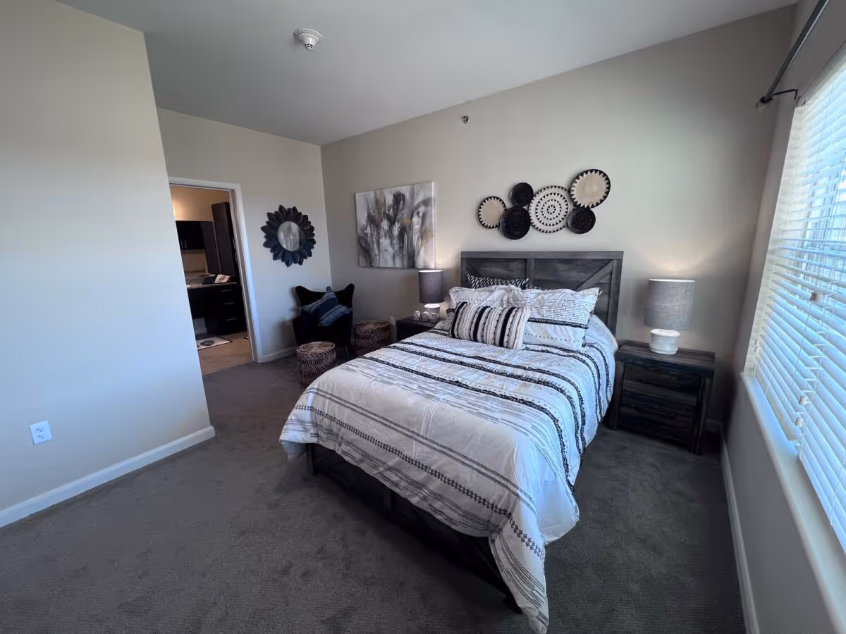 A bedroom with a neatly made bed featuring white and black striped bedding. There are two bedside tables with lamps on either side of the bed. The wall above the bed is decorated with circular woven baskets and a framed abstract painting. A black armchair with a pillow and two woven stools are placed near the corner. A window with blinds is on the right side, allowing natural light into the room. An open doorway leads to a bathroom area.