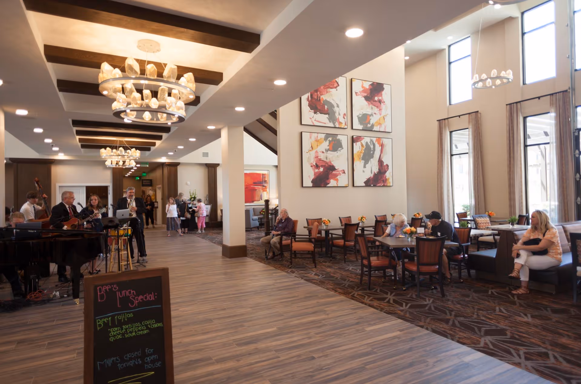 A spacious and well-lit common area in a senior living facility with wooden beams on the ceiling and modern chandeliers. On the left, a live band is performing with various instruments including a piano, guitar, saxophone, and drums. In the center and right, several seniors and visitors are seated at tables and chairs, some engaged in conversation. Large windows with curtains allow natural light to fill the room. Colorful abstract paintings hang on the wall above the seating area. A chalkboard sign in the foreground displays lunch specials.