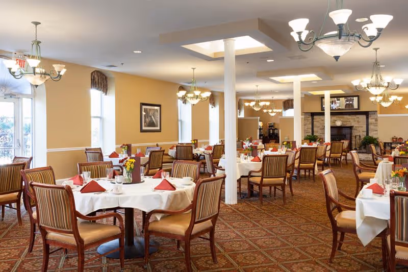 A spacious dining room with multiple round tables covered with white tablecloths, each set with red folded napkins, cups, and small flower arrangements. The room features beige walls, patterned carpet, several chandeliers, large windows with valances, and a fireplace in the background.