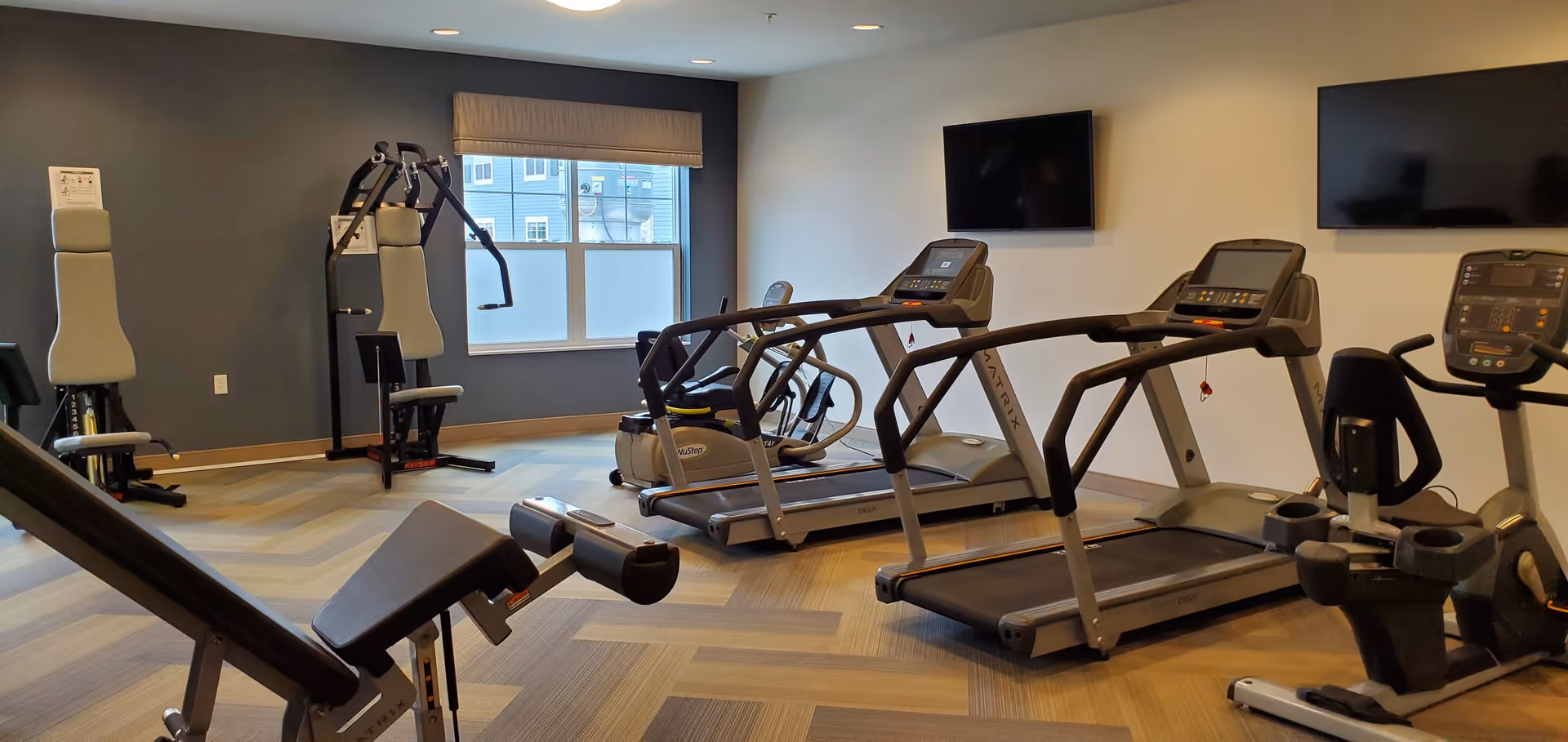 A fitness room with exercise equipment including treadmills, stationary bikes, and weight machines. The room has a large window with a beige valance, two wall-mounted flat-screen TVs, and a patterned carpet floor.